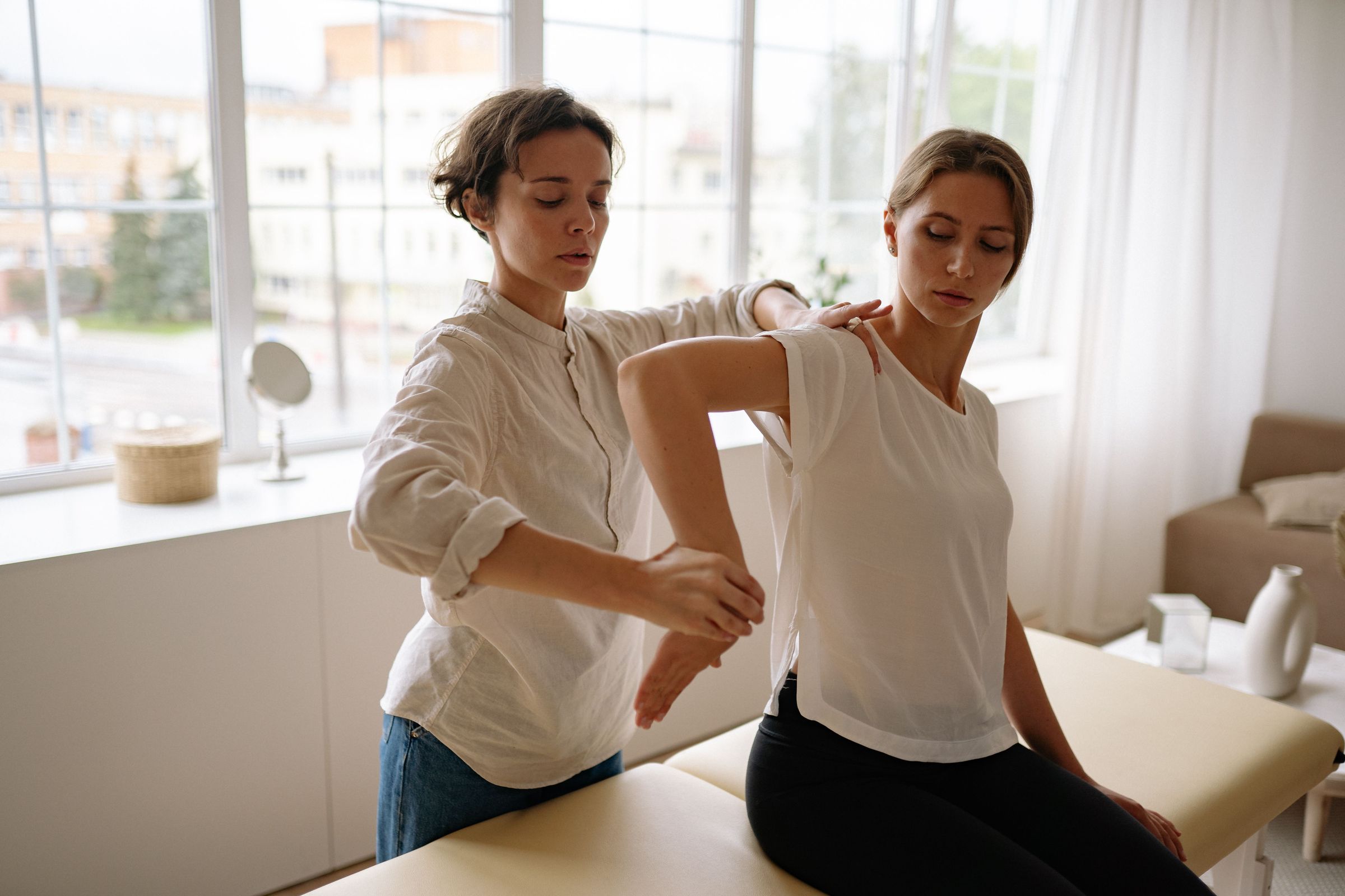 A physical therapist guiding a client through an arm stretch in a bright treatment room beside a large window
