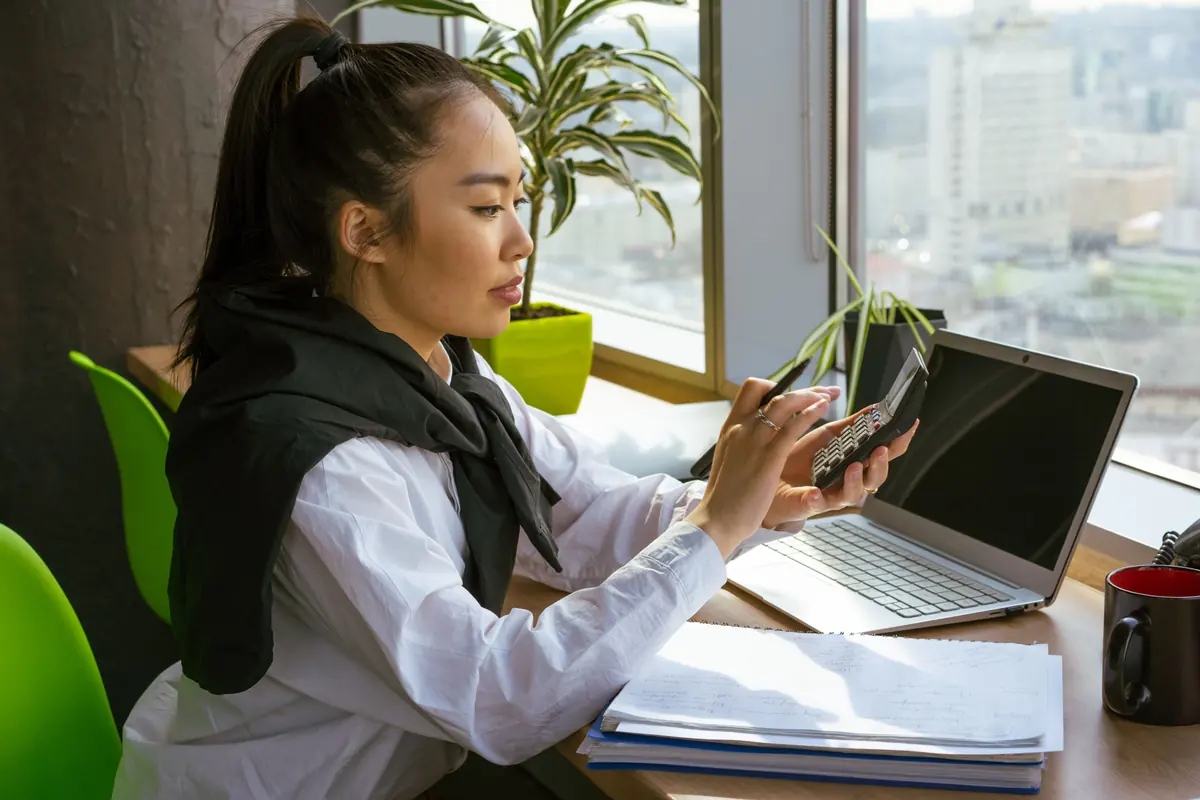 A woman at a window desk working a calculator beside an open laptop