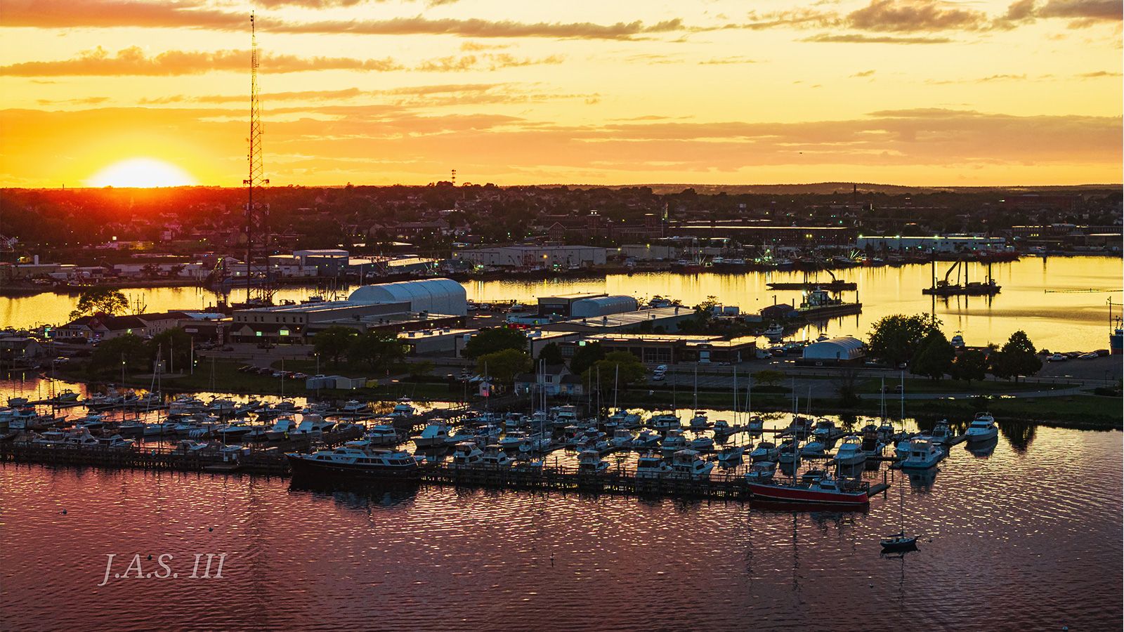 Sunset over a South Coast harbor, with a boatyard, work sheds and a marina full of boats on the near shore