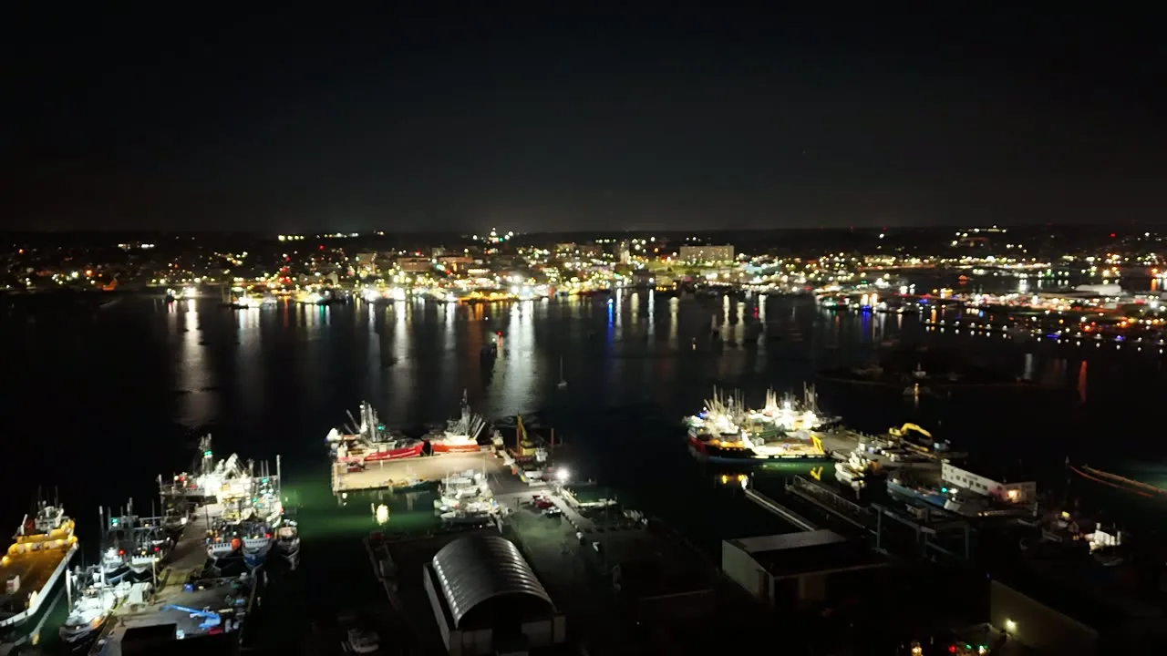 The working waterfront at night from above, with fishing boats lit at the docks and the city lights across the harbor