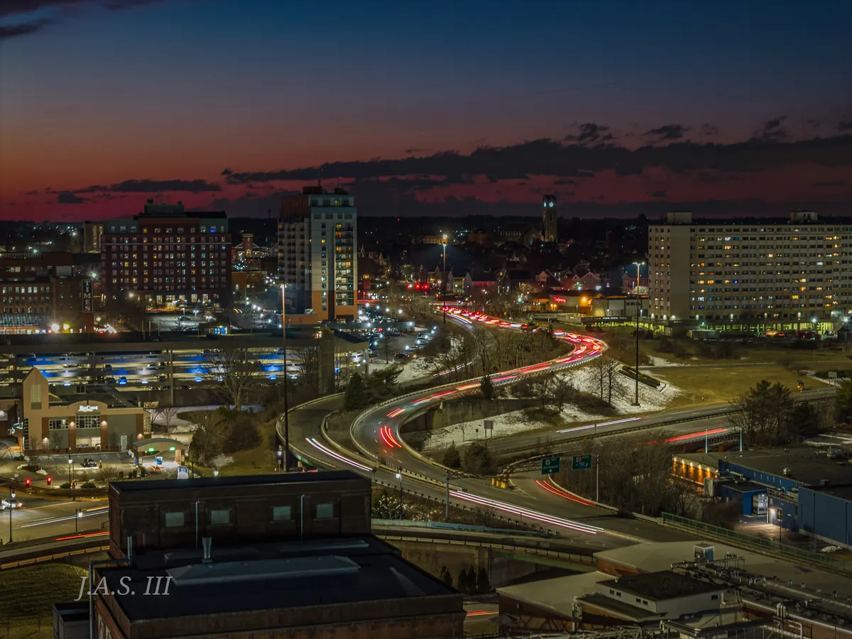 A South Coast city at night from above, with red and white traffic trails curving through the streets below a deep orange sky