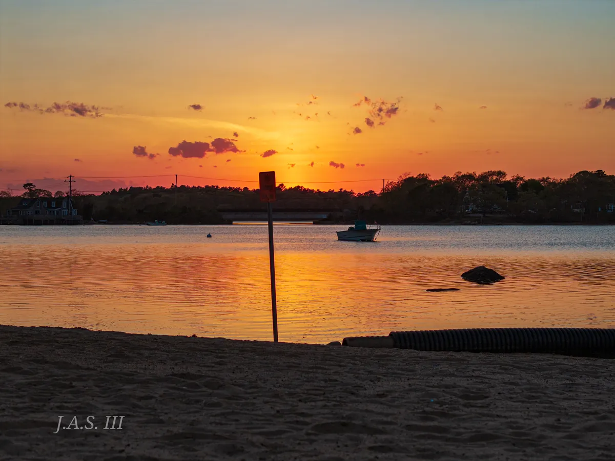 Sunset over a calm cove seen from a sandy beach, with a small boat on the glowing water