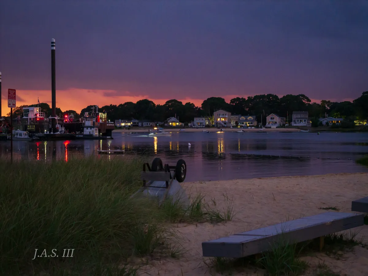 A harbor shore at dusk, with lit houses and a tall mast across the water and beach grass and a bench in the foreground